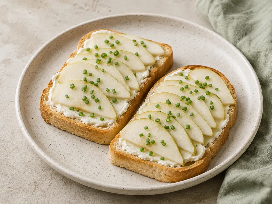 Zwei Scheiben helles Toastbrot mit Frischkäse, Birnenspalten und Schnittlauch auf einem hellen Teller